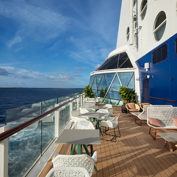 Cruise ship deck with white chairs and gray tables along glass railing overlooking the ocean under a blue sky