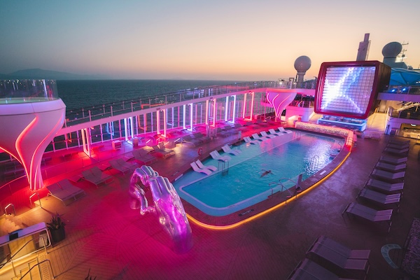 Cruise ship pool deck at sunset with pink neon lights, empty lounge chairs, and an illuminated sculpture near the pool edge.