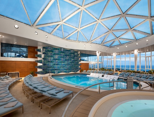 Luxurious indoor spa with a circular pool surrounded by rows of light blue lounge chairs and a glass skylight ceiling, letting in bright natural light.