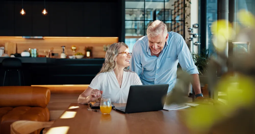 A couple smiling over a laptop at their kitchen table, researching and choosing a marriage retreat that fits them