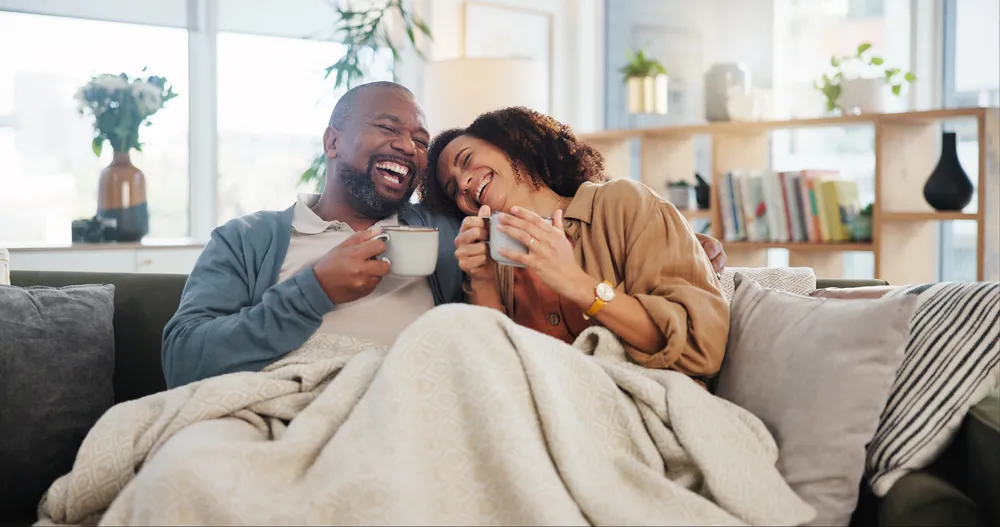 Happy couple laughing together under a blanket with coffee at home, relaxed and connected