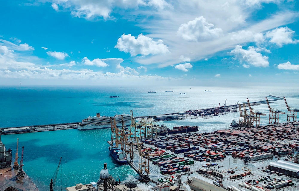 Port with rows of shipping containers, large cranes, and a cruise ship docked at the pier under a bright blue sky.